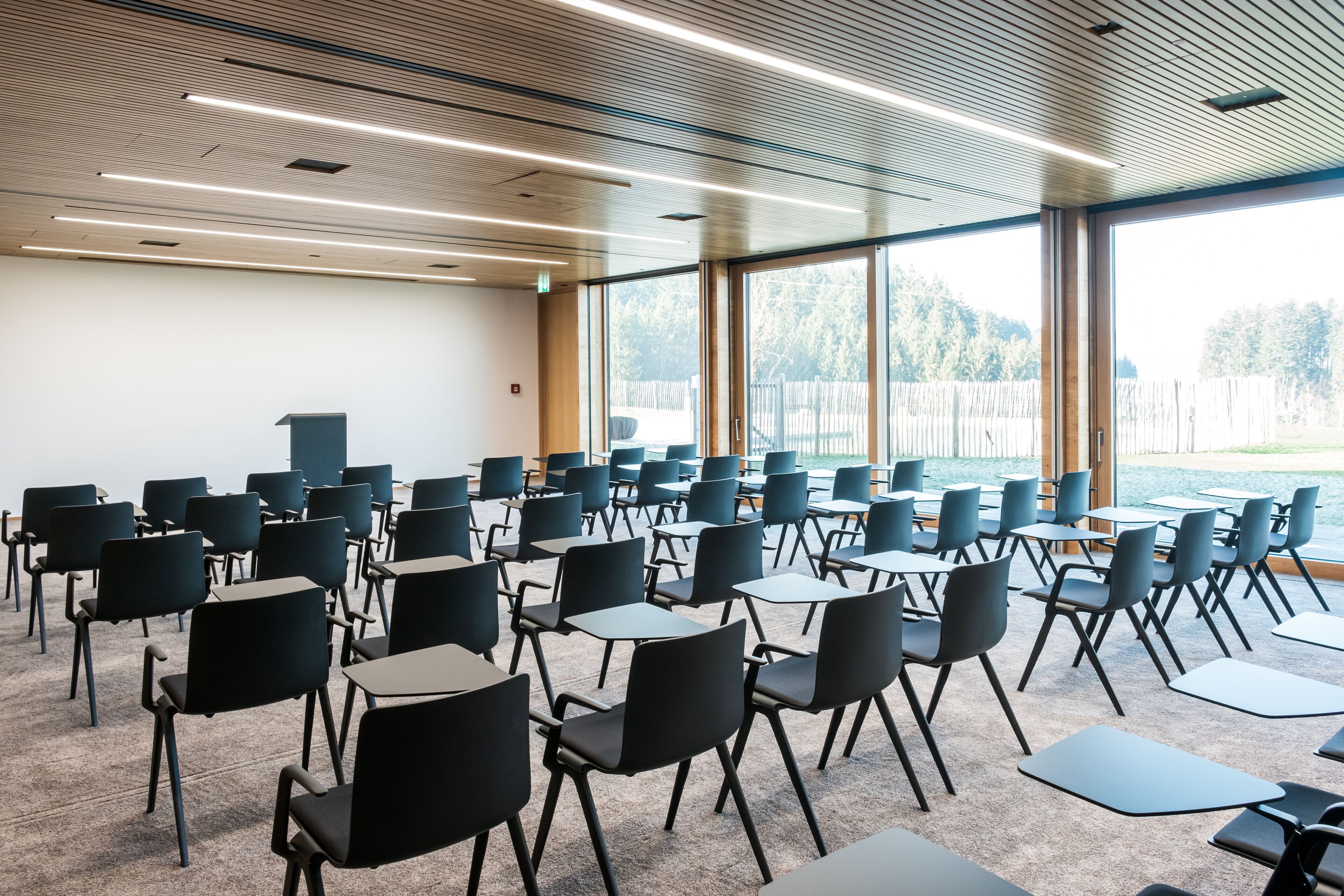 Un salón de conferencias con filas de sillas y mesas, ventanas amplias y techo de madera.