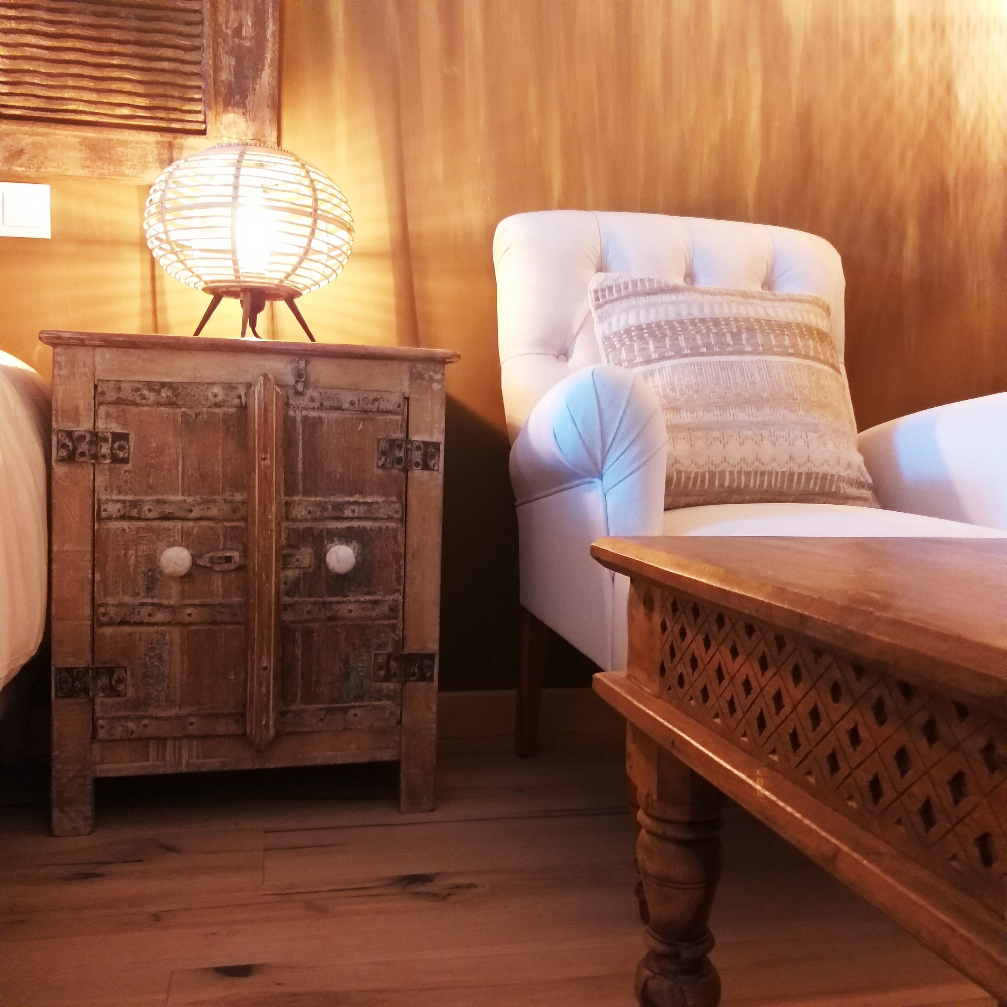 A wooden nightstand with a lamp, a white upholstered chair with patterned pillows, and a wooden table with a lattice design.
