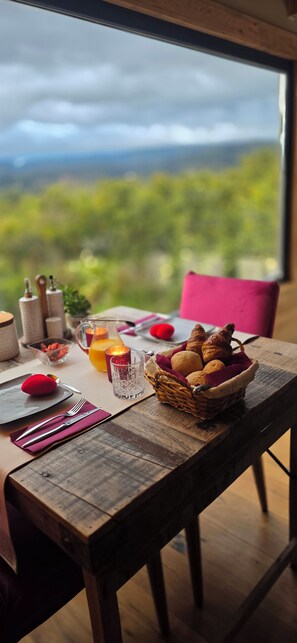 A dining table with a basket of bread, a glass of orange juice, and a red dessert on a wooden table with a view of a cloudy sky and greenery. - EL VIOLIN EXPERIENCIA (El Valle)