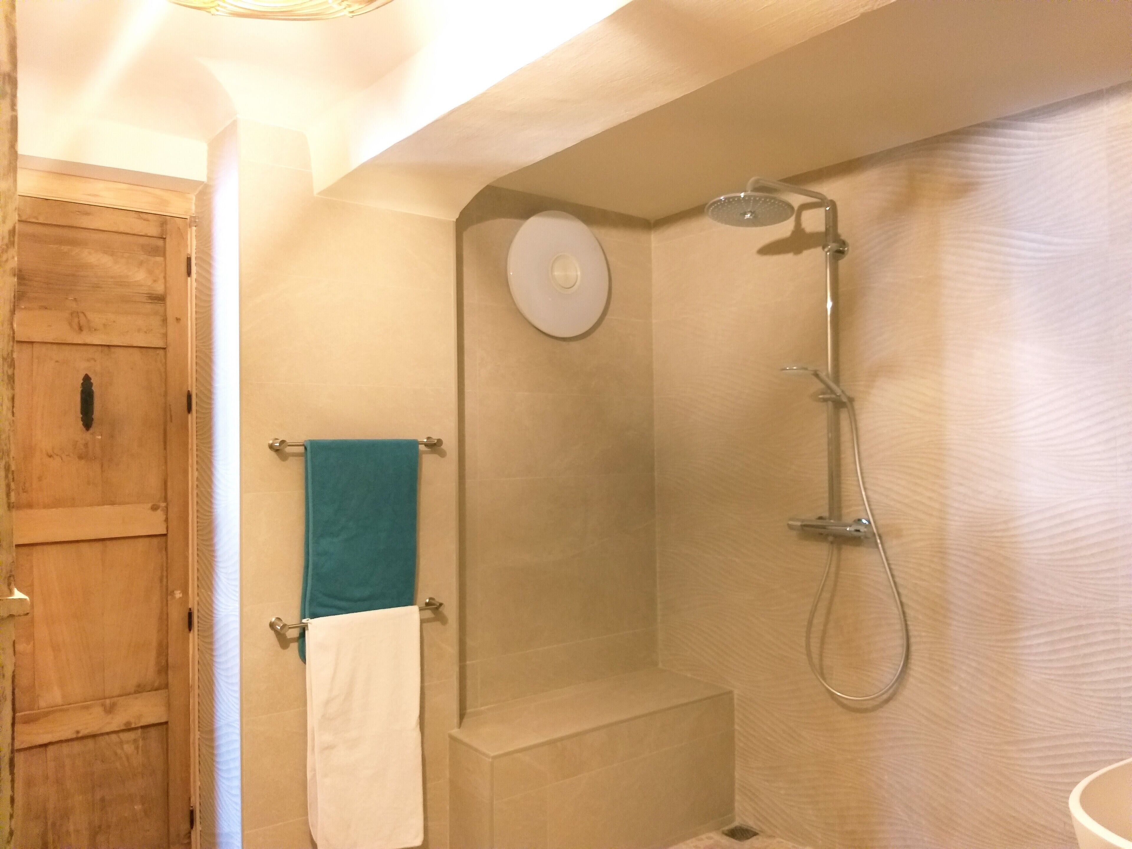 Un baño con puerta de madera, ducha con cabezal de ducha manual y un perchero con dos toallas.