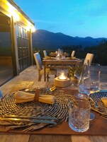 Una mesa al aire libre dispuesta para dos personas, con individuales tejidos, cubiertos y copas, un centro de mesa con velas y una vista de montañas al fondo.