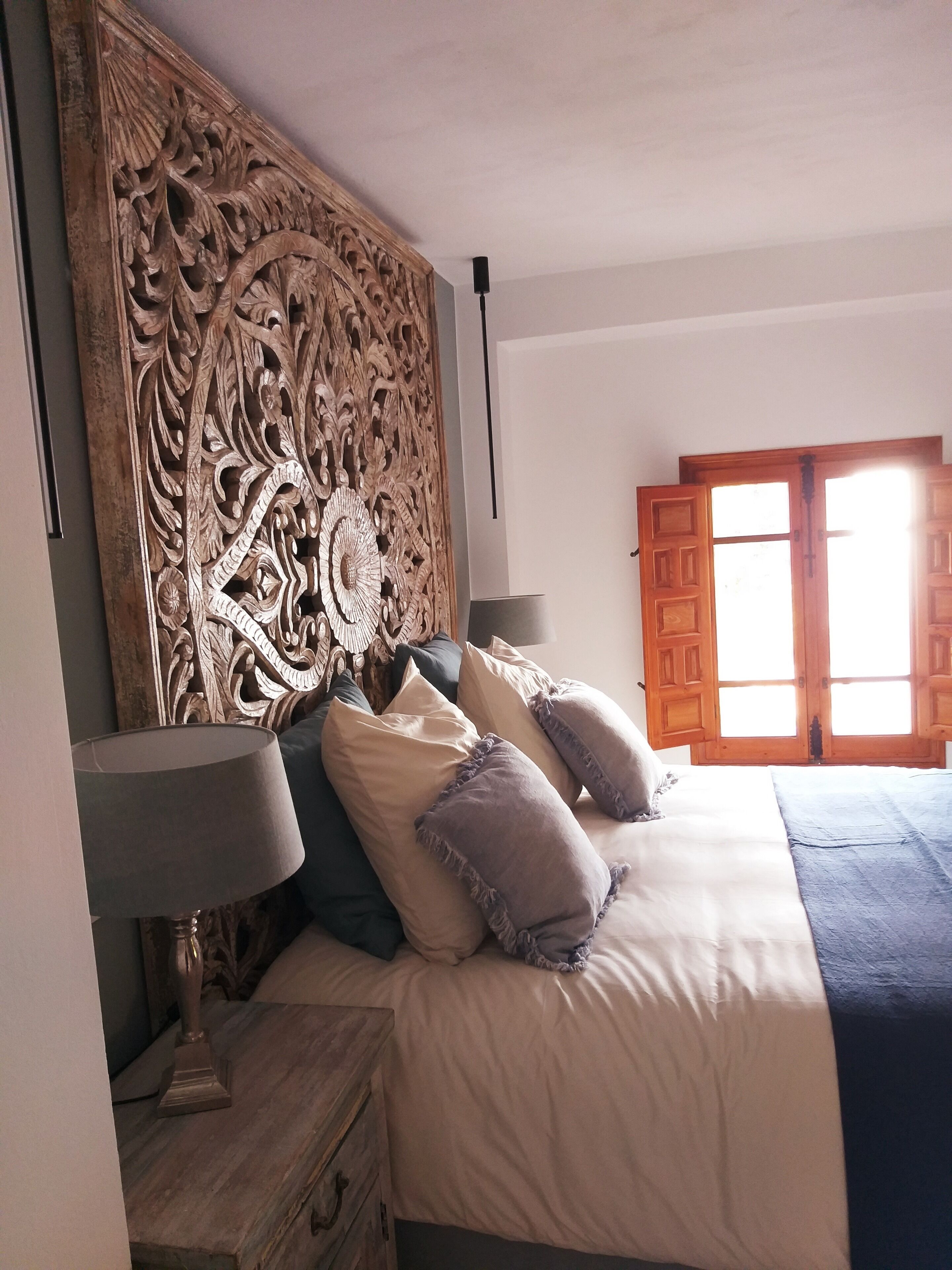 A bedroom with a large, ornate headboard, a bed with multiple pillows, a nightstand with a lamp, and a window with wooden shutters.