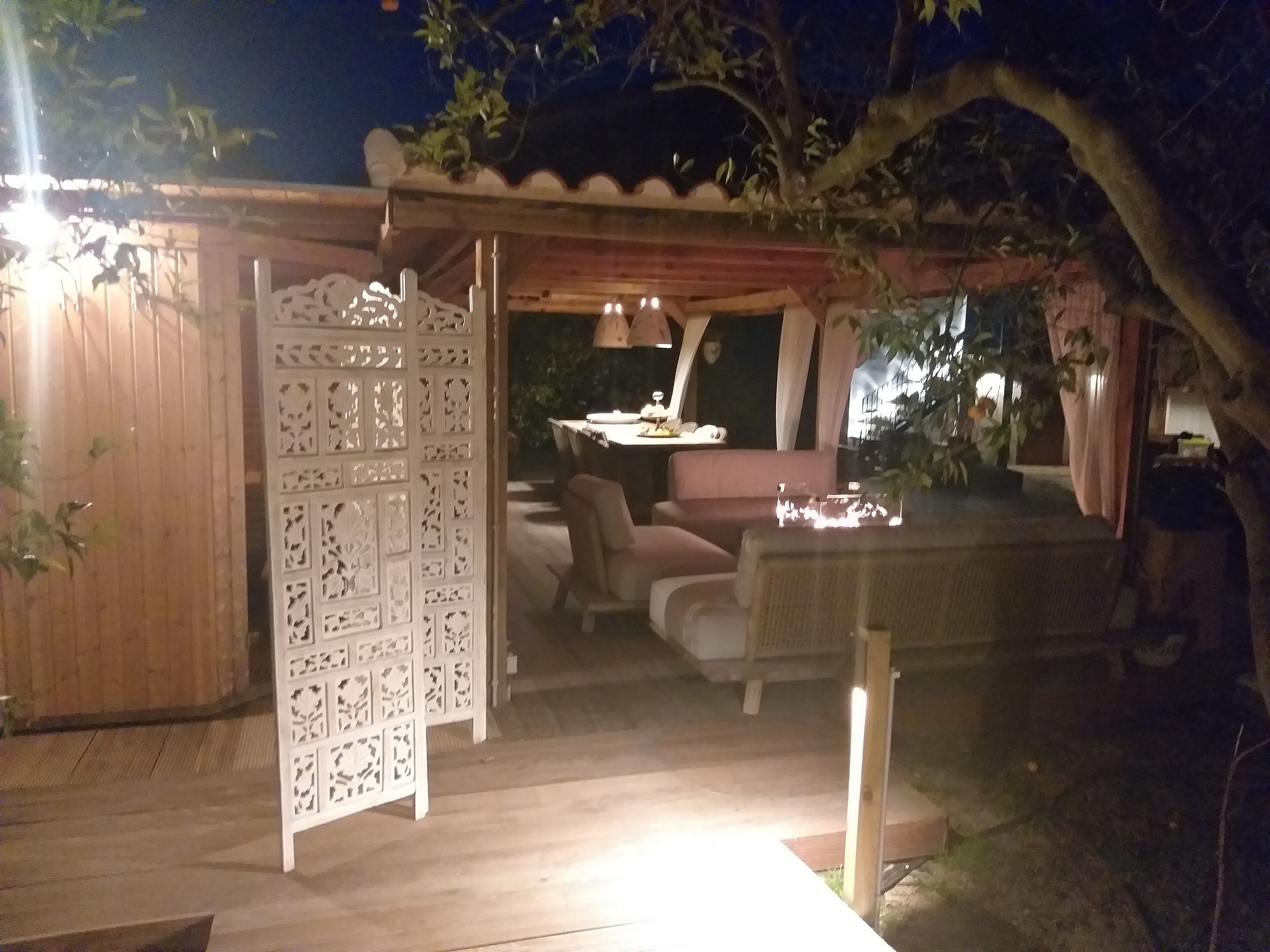 Un área de estar al aire libre con una mesa, sillas, un separador blanco decorativo y una pérgola con luces colgantes.