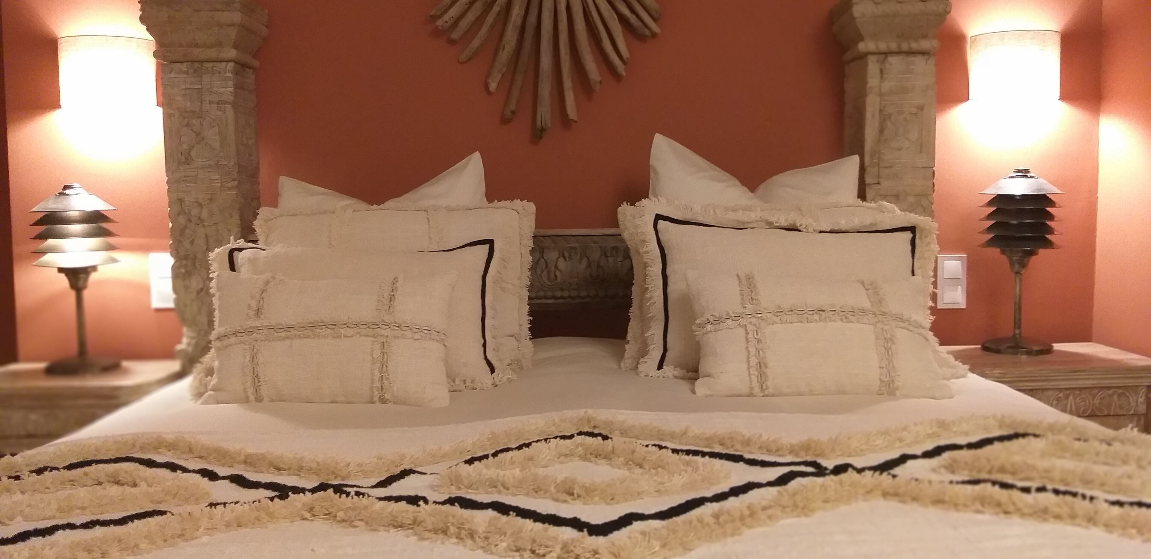 Una cama con múltiples almohadas y un tapete decorativo, iluminada por dos lámparas de pared.