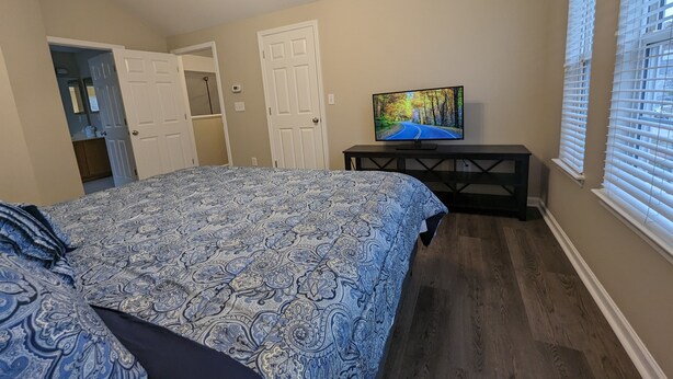 A bedroom with a bed, a TV on a stand, a door, and a window with blinds. - Cedar's Corner in Concord | Spacious & Clean (Concord)