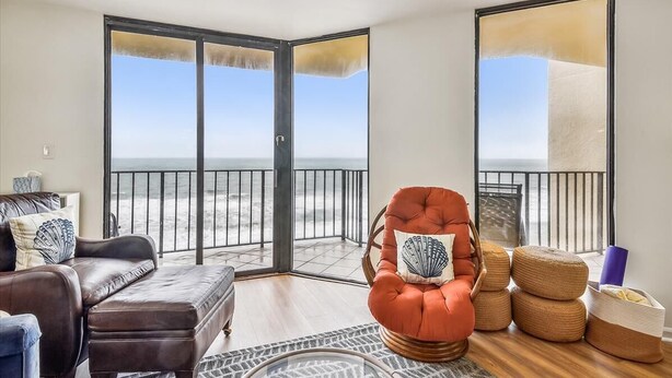 A living room with a view of the beach, a brown leather sofa, a red armchair with a seashell pillow, and wicker ottomans. - 10% off 3-7 nights thru Oct. A BEAUTY! Oceanfront 3 bedroom. Sleeps 6. Outdoor pool (North Myrtle Beach)