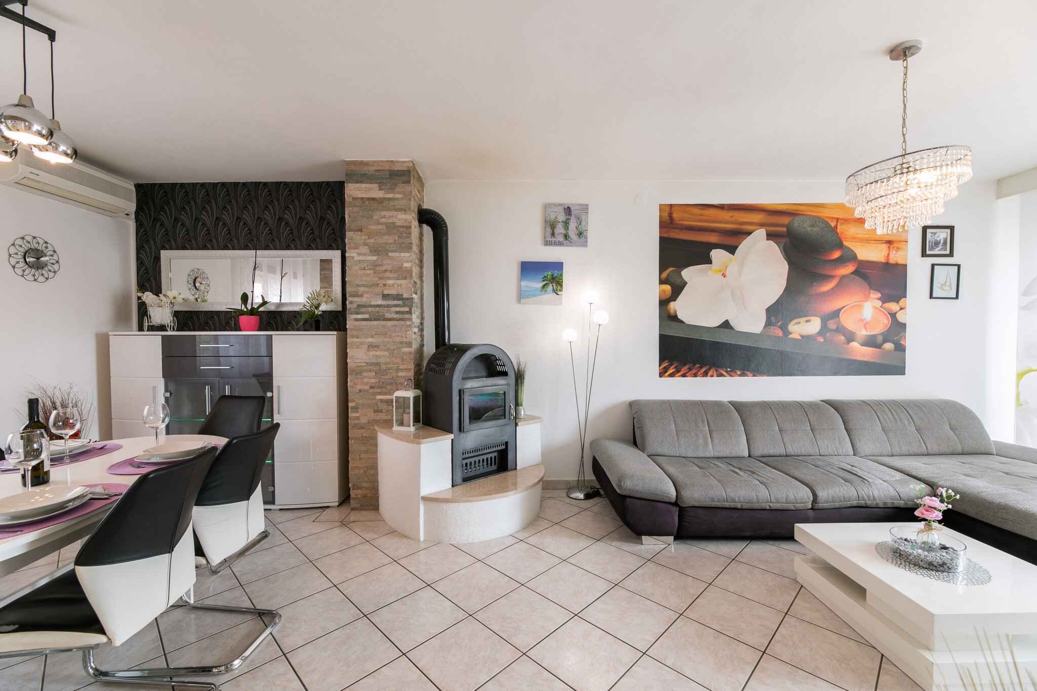 A modern living room with a grey sofa, a dining table with chairs, a fireplace, and a kitchen area with a built-in oven.