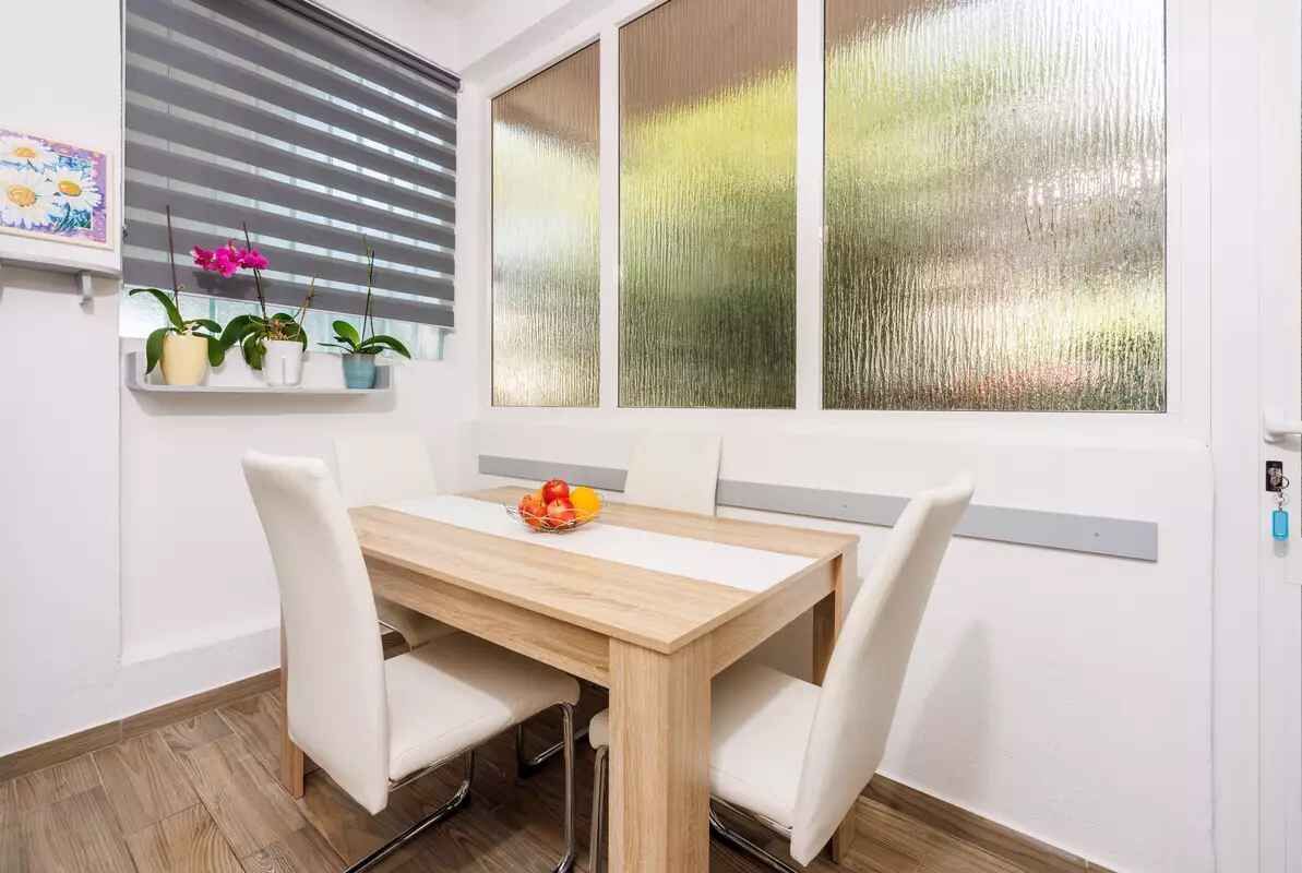 A dining area with a wooden table and white chairs, a window with blinds, and a shelf with potted plants.