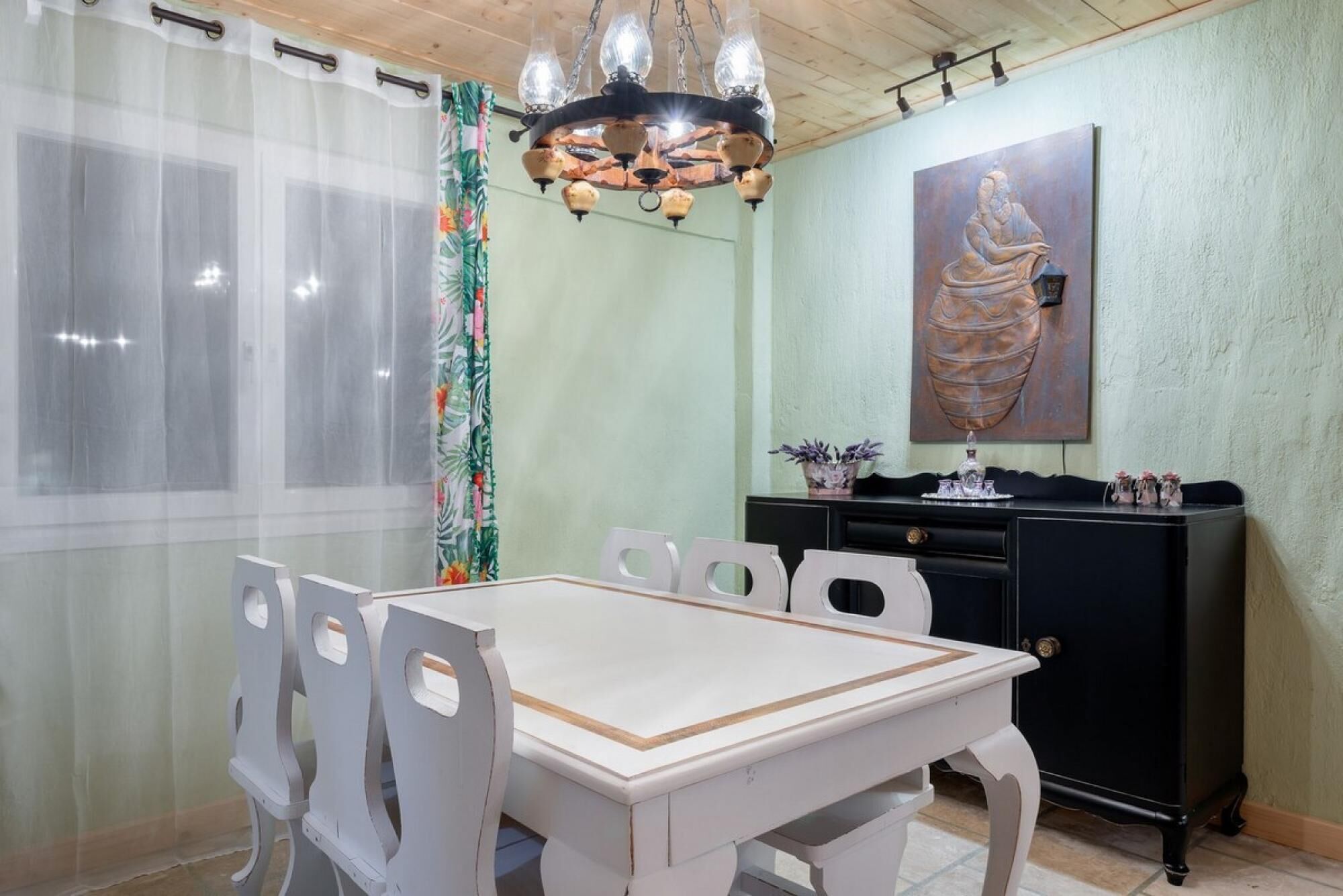 Een eethoek met een witte tafel en stoelen, een kroonluchter, een zwarte kast met decoratieve objecten en een schilderij aan de muur.