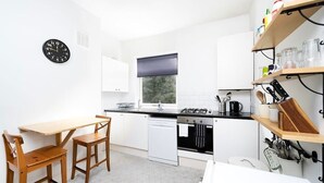 Een keuken met een eettafel, stoelen, een fornuis, een vaatwasser en een klok aan de muur.