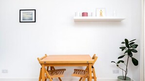 Een kamer met een houten tafel, twee stoelen, een witte muur, een ingelijste foto, een plank met spullen en een potplant.