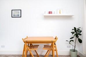 A room with a wooden table and two chairs, a white wall, a framed picture, a shelf with items, and a potted plant. - Lovely Top Floor Flat in Vibrant New Cross London (Greater London)