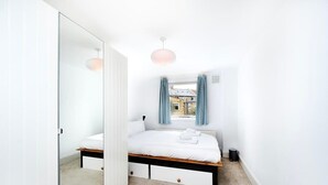 Un dormitorio con cama, mesitas de noche, ventana con cortinas, radiador y puerta que da a otra habitación.