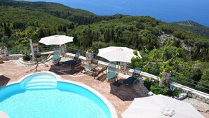A pool area with lounge chairs and umbrellas, surrounded by lush greenery and a view of the sea. - Villa Fantasia Kefalonia (Kefalonia)