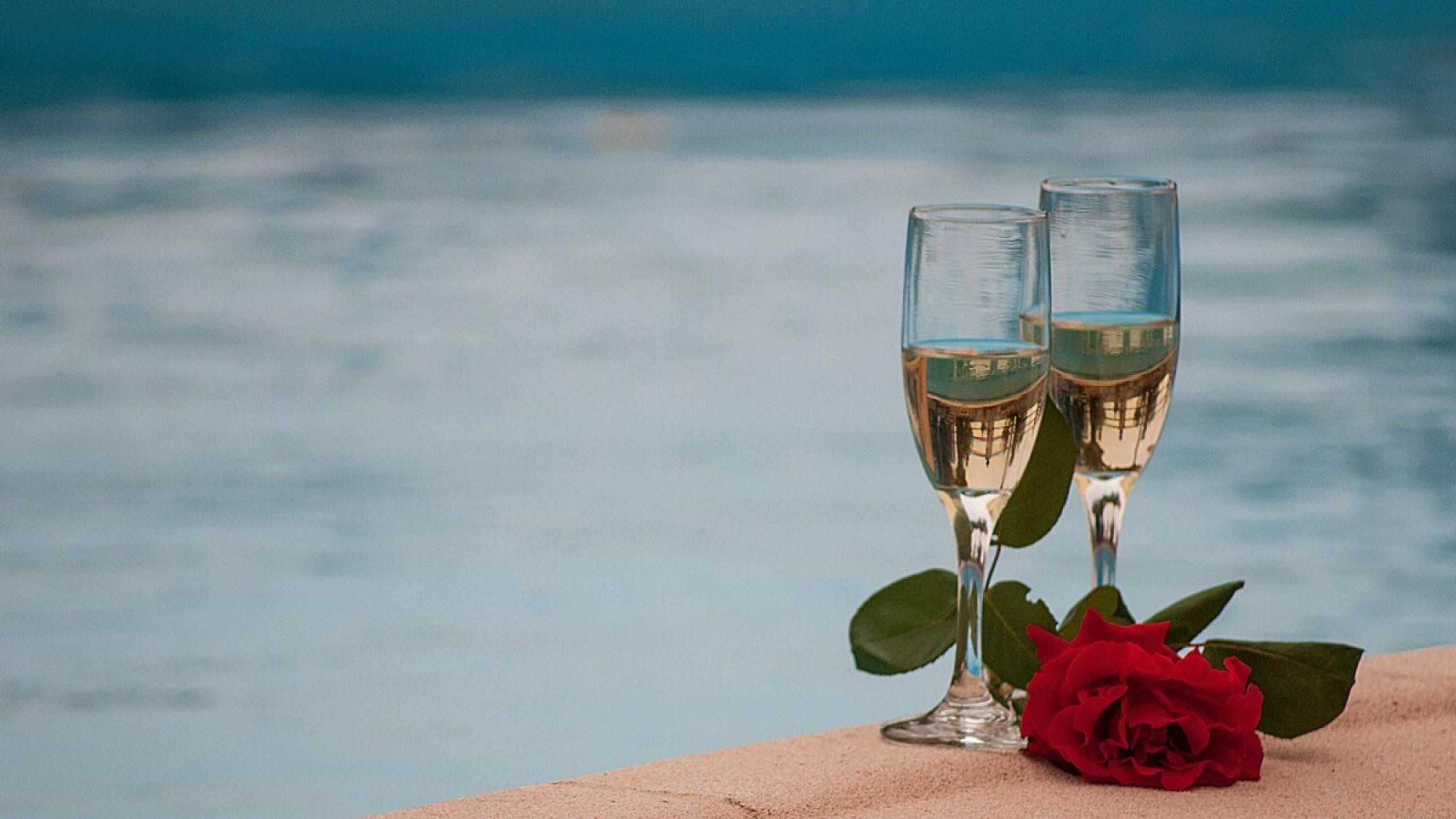 Two champagne flutes with a red rose beside them, placed on a surface with a blurred body of water in the background.