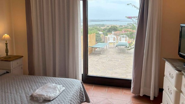 A bedroom with a bed, a nightstand with a lamp, a dresser, and a large window with curtains. - Holiday home with private pool and sea view (Punta de Su Turrione)