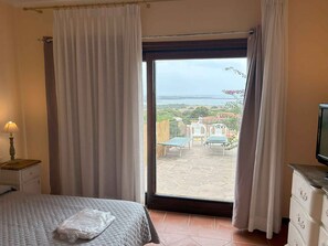 A bedroom with a bed, a nightstand with a lamp, a dresser, and a large window with curtains. - Holiday home with private pool and sea view (Punta de Su Turrione)