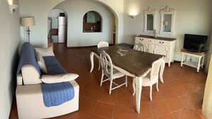 A living room with a sofa, dining table, chairs, a TV, and a kitchen visible through an archway. - Holiday home with private pool and sea view (Punta de Su Turrione)