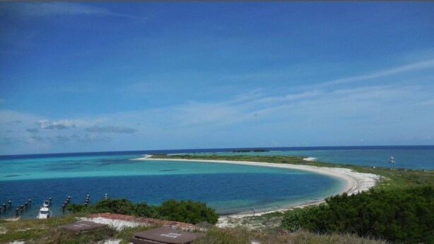 Un paisaje costero con aguas turquesas cristalinas, una playa arenosa y un faro a lo lejos.
