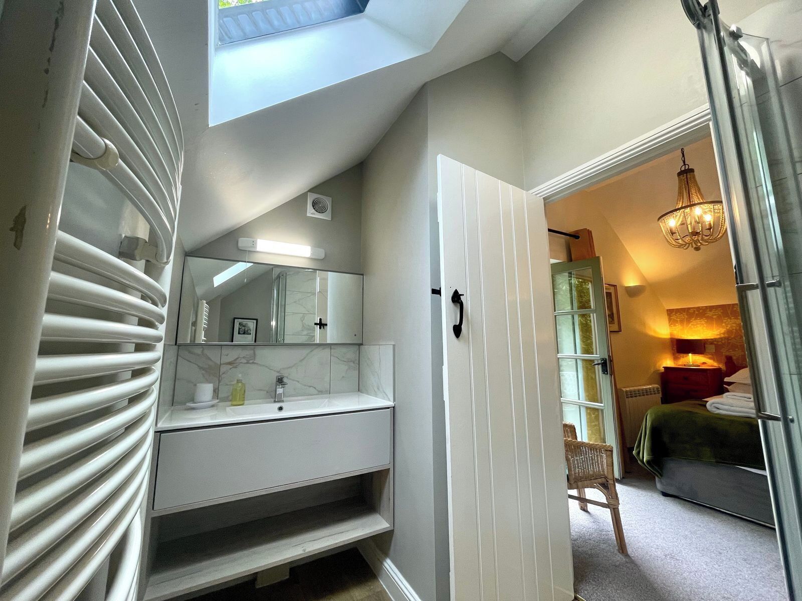 Une salle de bain moderne avec un puits de lumière, un grand miroir, un meuble-lavabo blanc et une porte donnant sur une chambre à coucher équipée d’un lit et d’un lustre.