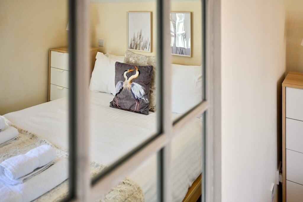 A neatly made bed with white linens, a pillow with a bird design, and a bedside table with a lamp.