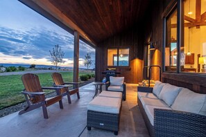 A covered outdoor seating area with chairs and a sofa, a grill, and a view of the landscape.