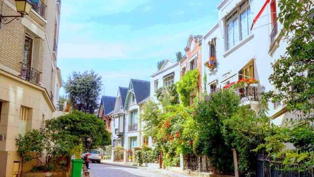A narrow cobblestone street lined with traditional European-style buildings, featuring balconies with hanging flower baskets and lush greenery. - Seb's Place Montmartre | Paris Safe and quiet area (Paris)