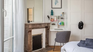 A bedroom with a fireplace, a grey chair, a bed, a white wardrobe, and a shelf with various items. - Seb's Place Montmartre | Paris Safe and quiet area (Paris)