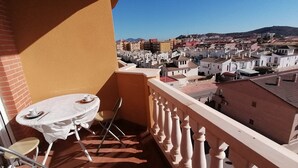 Un balcon avec une table et une chaise, donnant sur une zone urbaine densément peuplée, avec divers bâtiments et une colline visible au loin.