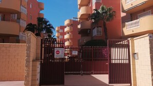 Une entrée fermée par un portail donnant accès à un immeuble résidentiel doté de nombreux balcons, avec un ciel bleu dégagé.