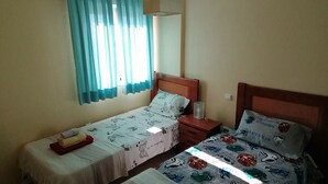 Une chambre avec deux lits simples, chacun recouvert d’une courtepointe blanche et agrémenté d’un oreiller. Une table de chevet avec une lampe se trouve entre les deux lits. Les murs de la chambre sont de couleur beige et il y a une fenêtre avec des rideaux bleus.