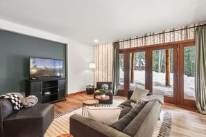 A modern living room with a large flat-screen TV, dark leather sofas, a wooden coffee table, and a view of a snowy landscape through glass doors.