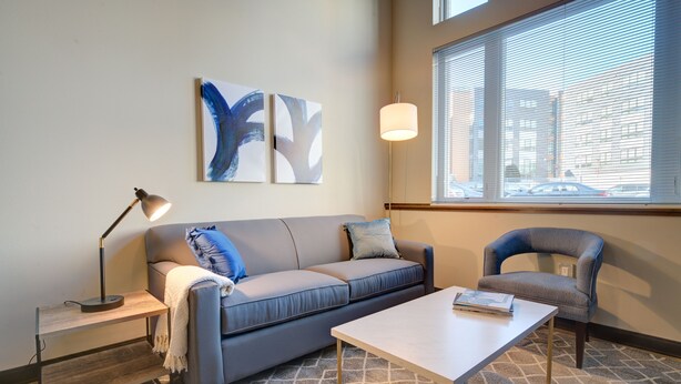 Ein modernes Wohnzimmer mit einer Couch, einem Couchtisch, einem Beistelltisch und einem Sessel. An der Wand hängen abstrakte Gemälde und ein Fenster mit Jalousien.