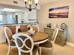 A dining area with a round table and chairs, a kitchen in the background, and a sunset painting on the wall.
