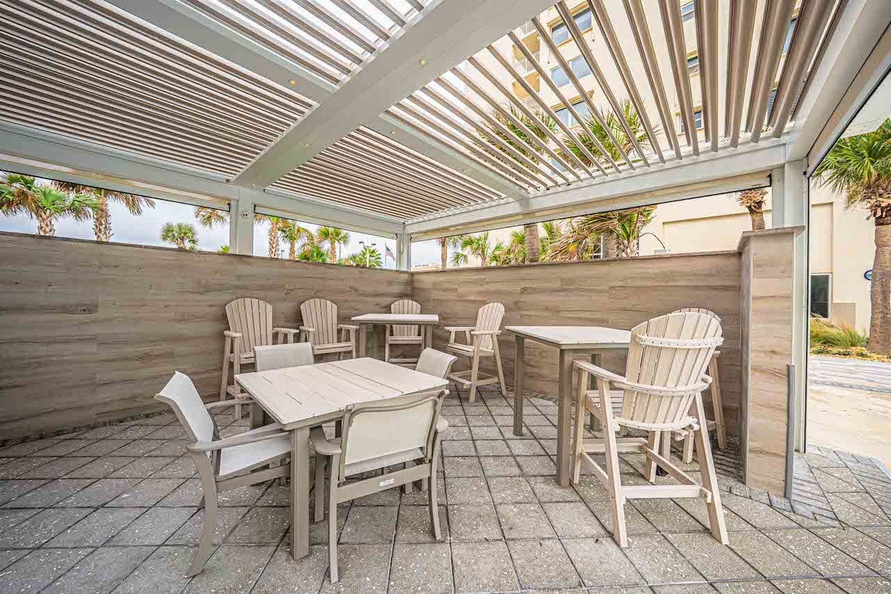 An outdoor patio with wooden tables and chairs, a pergola overhead, and a view of palm trees and a building in the background.