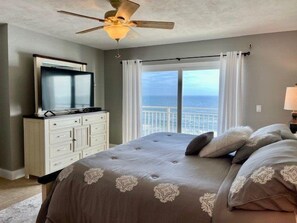 A bedroom with a bed, a television on a dresser, a ceiling fan, and a view of the ocean through large windows.