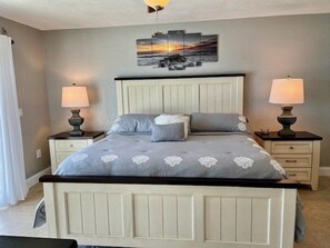 Un dormitorio con una cama, dos mesitas de noche, un velador, una obra de arte en la pared y un ventilador de techo.