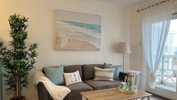 Un salón con un sofá gris, una mesa de centro de madera, un cuadro grande de una playa, una planta en maceta y vistas al exterior a través de una ventana con cortinas de encaje.