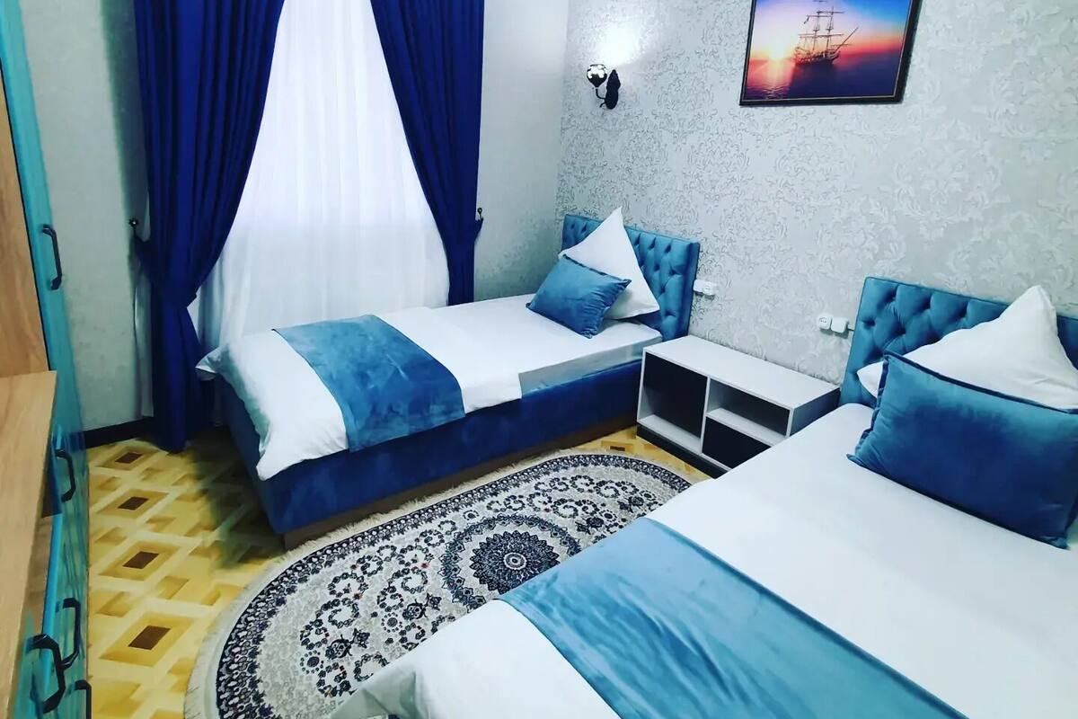 Une chambre d’hôtel avec deux lits, une grande fenêtre avec des rideaux bleus, un tapis à motifs et un tableau accroché au mur.