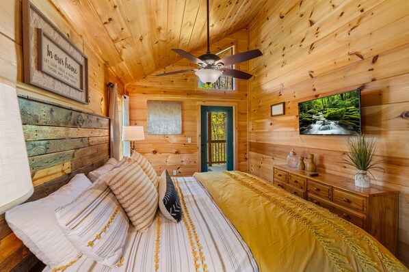 Un dormitorio de cabaña de madera acogedor, con una cama, un ventilador de techo, un televisor y una puerta que da a un balcón.