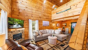 Acogedor interior de cabaña de madera con chimenea de piedra, sofá modular, comedor y televisor montado en la pared.