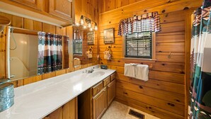 A cabin-style bathroom with wooden walls, a large mirror, a sink with a white countertop, and a window with a plaid curtain. - Always Together Forever: Make Memories That Last (Sevierville)