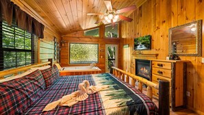 A cozy cabin interior with a large bed, a fireplace, a television, and a ceiling fan. - Always Together Forever: Make Memories That Last (Sevierville)