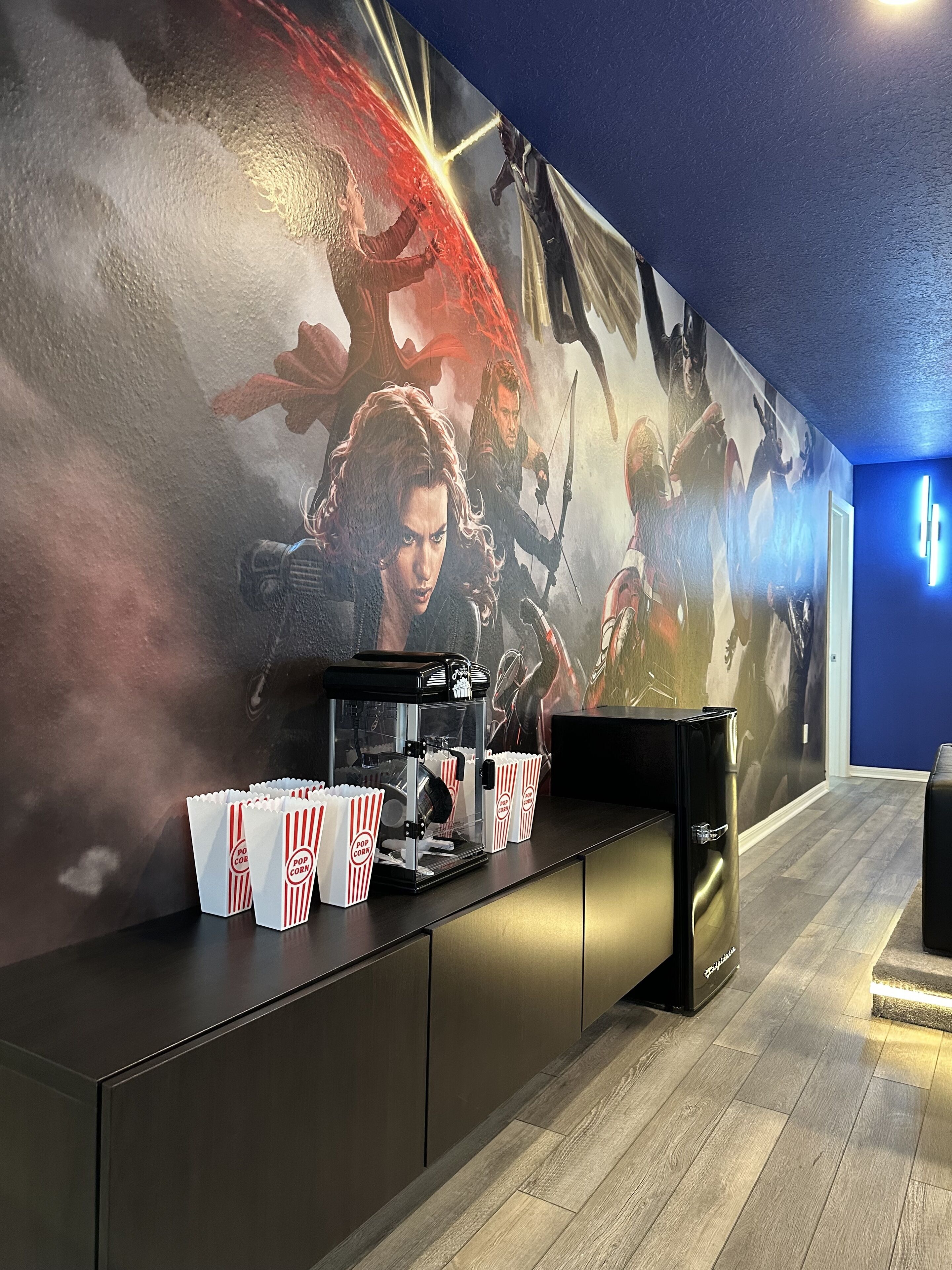 Ein Kino-Foyer mit einem großen Filmplakat an der Wand, einer Popcornmaschine und einer schwarzen Sitzgruppe.