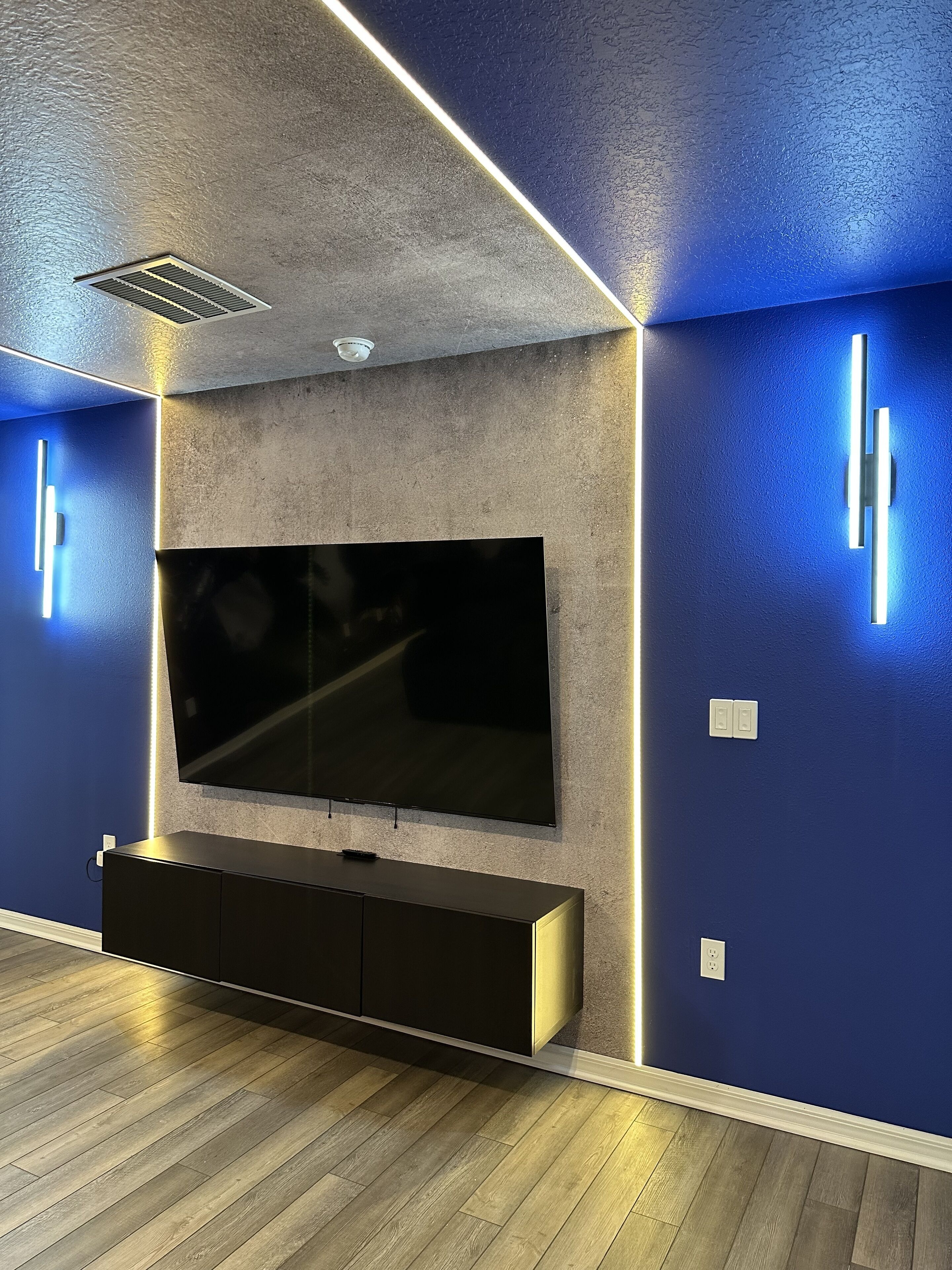Una sala de estar moderna con un televisor de pantalla plana grande montado en una pared texturizada, un mueble empotrado debajo y iluminación de acento azul a lo largo de los bordes.