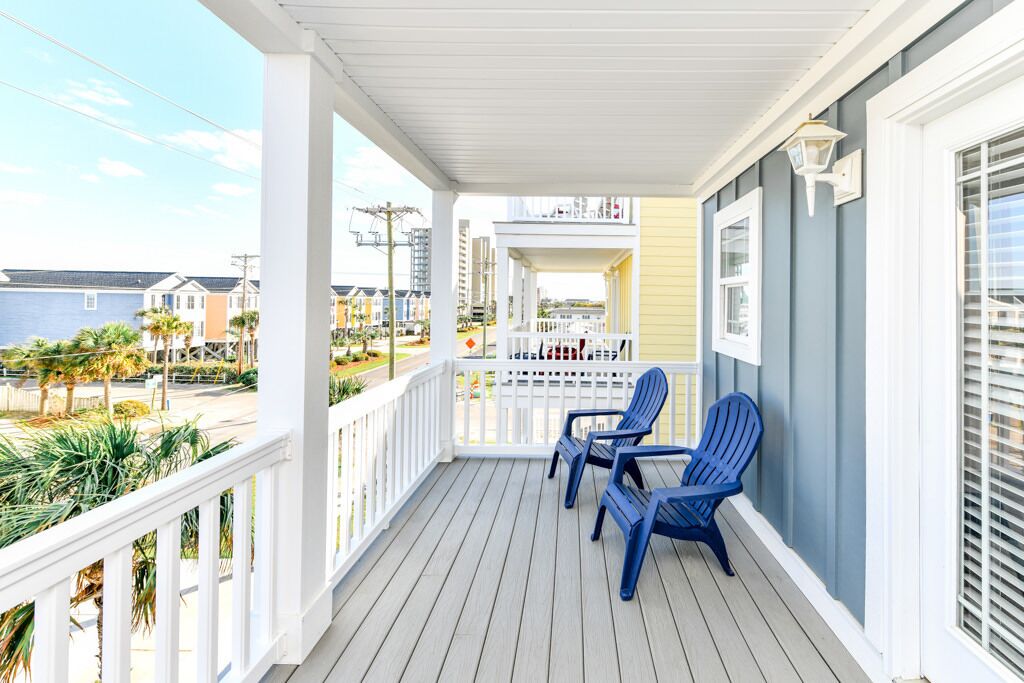 Une véranda couverte avec des balustrades blanches, deux fauteuils Adirondack bleus et une vue sur la plage et les maisons voisines.