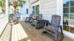 A wooden deck with outdoor furniture, including chairs and a table, and a white building with large windows in the background. - 4 BR 2nd Row Beach House with a Private Pool! Beachy Keen (Surfside Beach)
