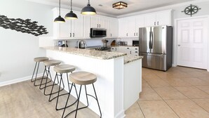 A modern kitchen with white cabinets, a stainless steel refrigerator, a microwave, and a granite countertop. There are four bar stools and pendant lights above the island.