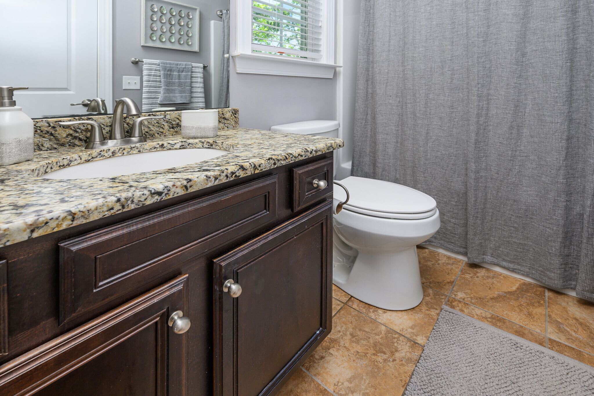 A bathroom with a granite countertop sink, dark wood cabinets, a white toilet, and a window with blinds.