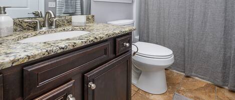 A bathroom with a granite countertop sink, dark wood cabinets, a white toilet, and a window with blinds.